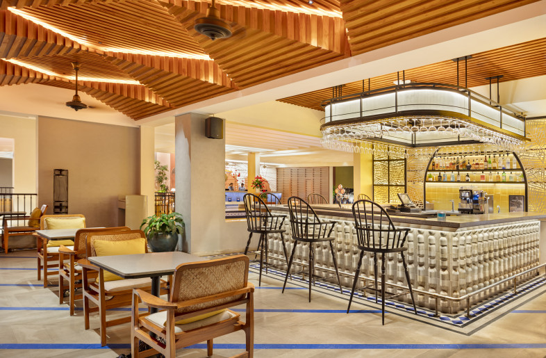 Taverna Bar dressed in custom cast concrete tiles and a blue grey cast in situ  terrazzo counter top. Yellow glazed tiles and backlit palm motif screens set off the bar and provide a vibrant display. The bar  framed through an opening in the wall, is visible from the lobby . The ceiling with its faceted fluted wood details recalls memories of shacks at the beaches in Goa. - Andre Fanthome (Studio Noughts and Crosses)