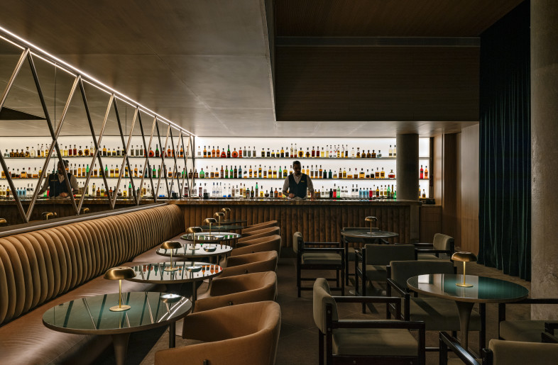SARAU BAR at PULSO HOTEL | The cozy venue hosts both pocket shows and listening bar nights, offering signature cocktails and small bites. Defined by wood-acoustic lowered ceilings, bronze mirrored walls, a marble bar, concrete pillars, and carpeted flooring, the space exudes a warm and intimate atmosphere. An open performance area is integrated into the layout, where soft, ambient lighting enhances the live music experience. - Fran Parente