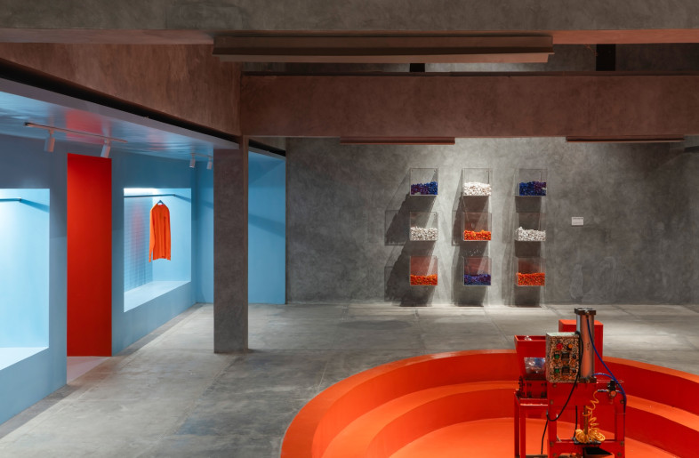 A central, tangerine-hued conversation pit anchors the space, surrounded by plastic recycling machines and upcycling stations. - Ernest Theofilus