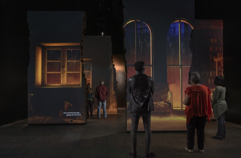 The 1921 Tulsa Race Massacre: Visitors immediately step into the cacophony of the 1921 Tulsa Race Massacre. Projected onto multiple surfaces, environmental motion graphics immerse visitors in the destruction and violence of the night, while they listen to the recorded memories of survivors. The back sides of the projection surfaces offer insight into experiences of different community members like the African Blood Brotherhood and white witnesses. - Local Projects/ Mel Willis