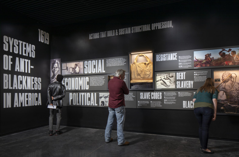 Systems of Anti-Blackness: Monumental imagery and embedded casework make plain the political, economic, and social systems of anti-Blackness that created the conditions for the 1921 Tulsa Race Massacre. Events are focused on Tulsa but framed in the national context and key features include artifacts of control and violence like slave shackles and a robe from the domestic terrorist organization, the Ku Klux Klan.  The final wall in the Systems of Anti-Blackness space chronologically catalogues the events leading up to and the hours of the 1921 Tulsa Race Massacre. - Local Projects/ Mel Willis