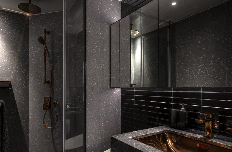 A moody en-suite bathroom outfitted in light grey terrazzo and dark chrome tiles paired with a copper basin and fixtures. - Mr. Edmon Leong